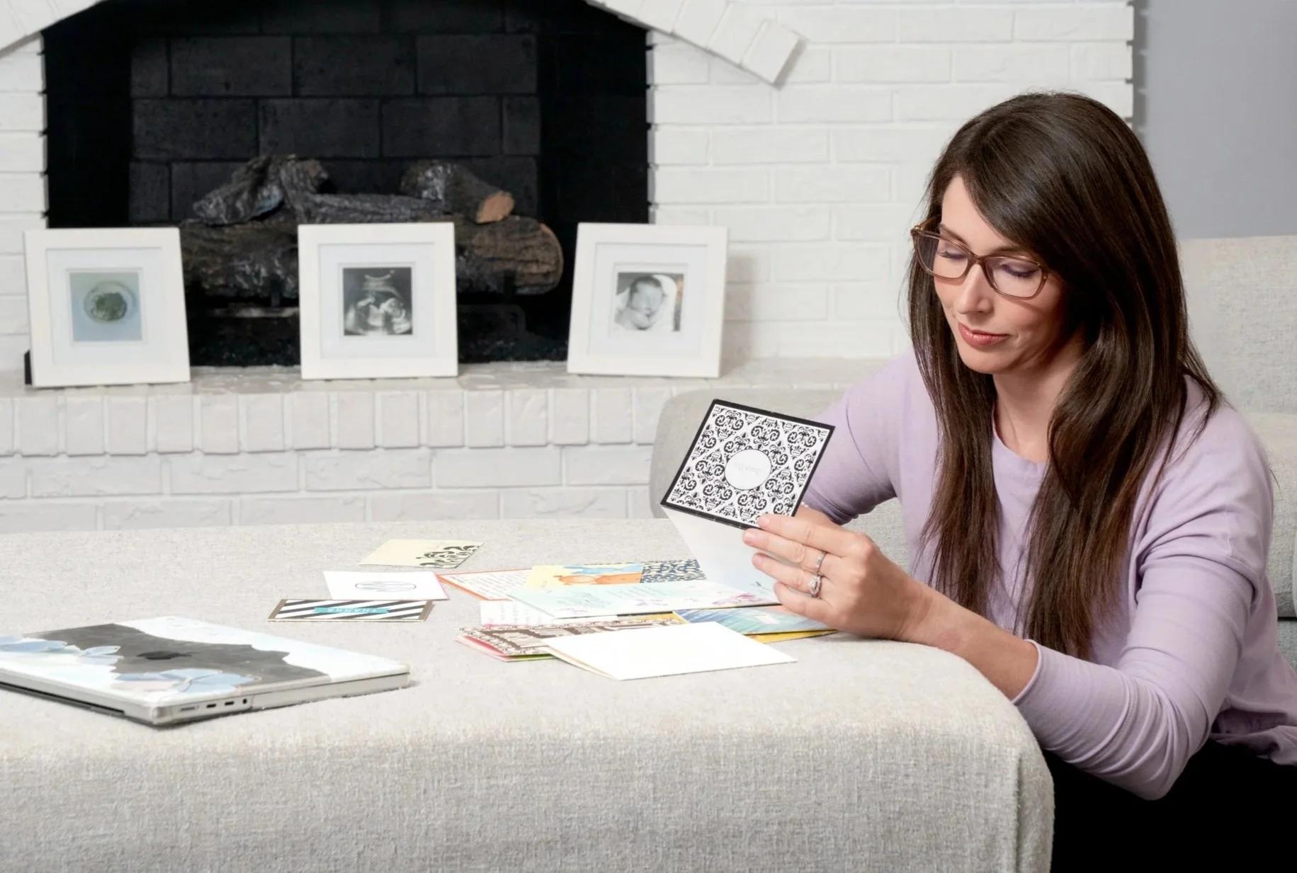 Woman sitting at a table looking at a card, with various other cards and a book on the table. Behind her, a white brick fireplace with three framed photos and firewood inside.