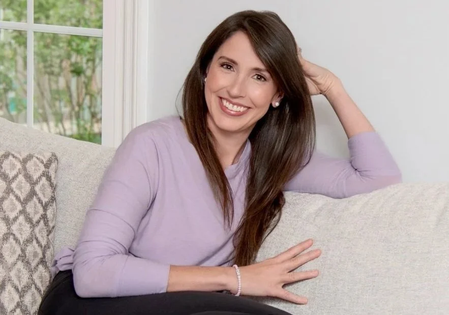 A woman with long brown hair smiling, sitting on a beige couch in a living room with a window behind her.