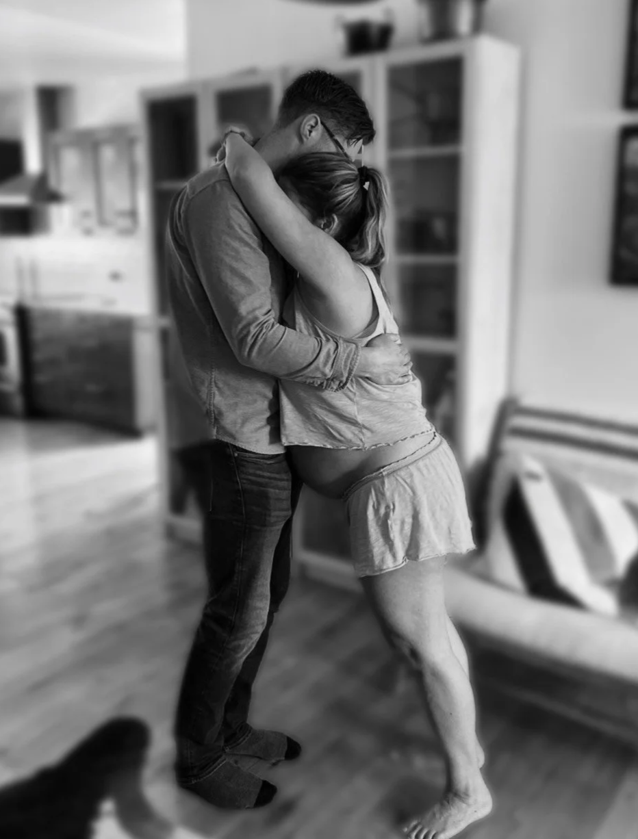A couple hugging in a living room, with the woman appearing pregnant, in black and white.
