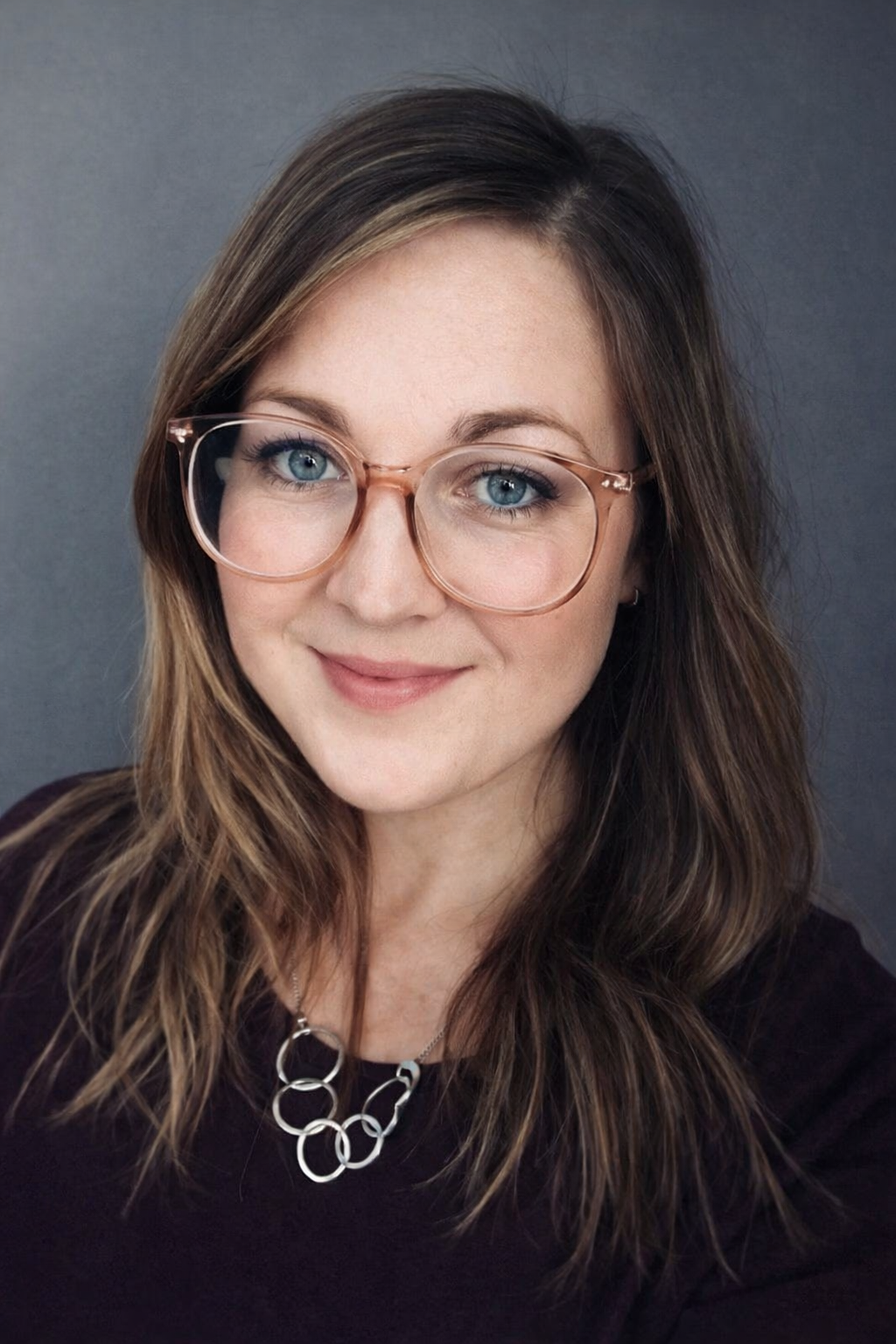 A woman with shoulder-length brown hair, wearing large round glasses with a clear frame, smiling at the camera against a plain background. She is wearing a black top and a silver necklace with interlocking rings.