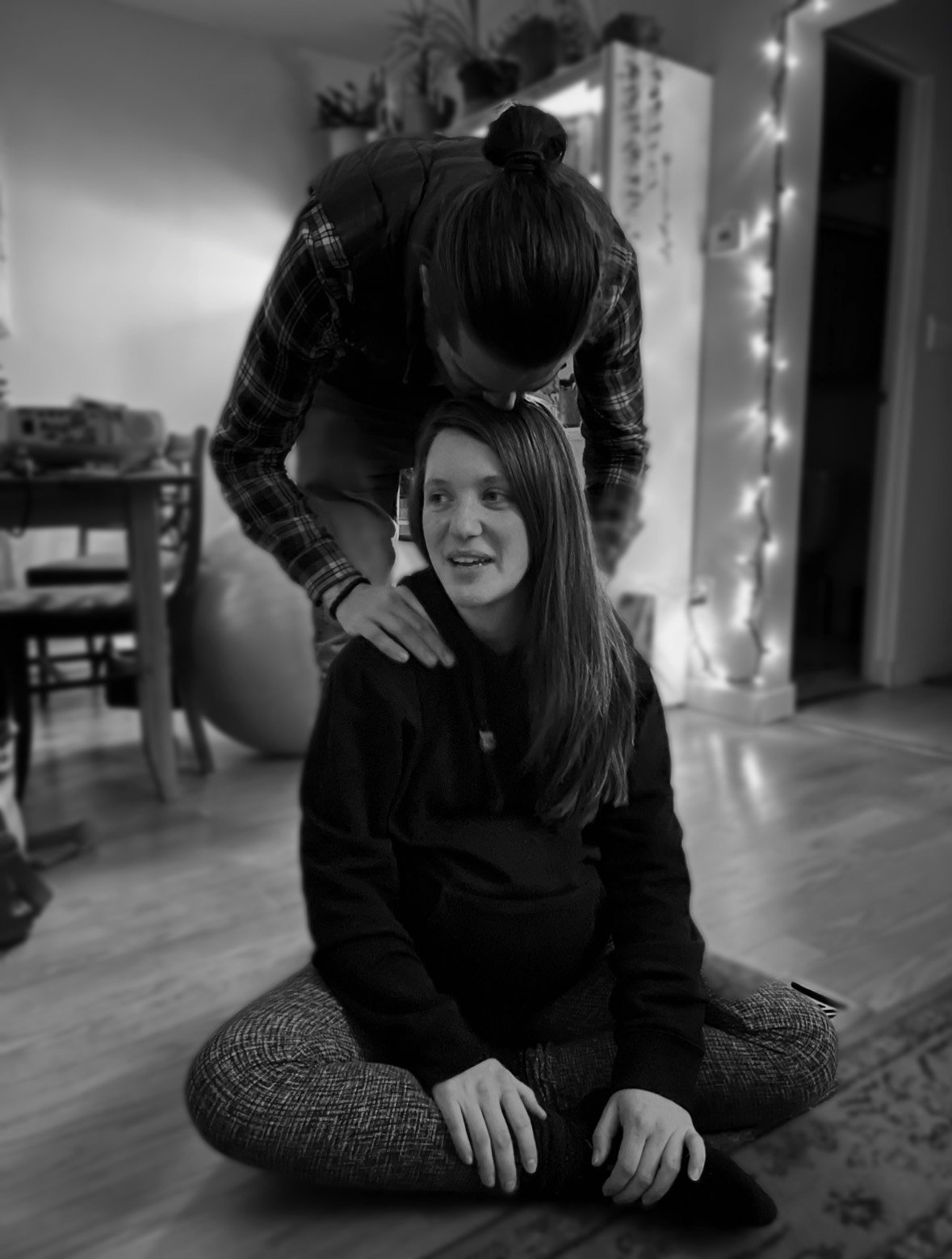 A black and white photo of two women, one sitting cross-legged on the floor and the other leaning over her, touching her shoulder. The woman sitting appears relaxed, while the woman leaning over looks engaged. The background features a decorated living space with chairs, a table, plants, and string lights.