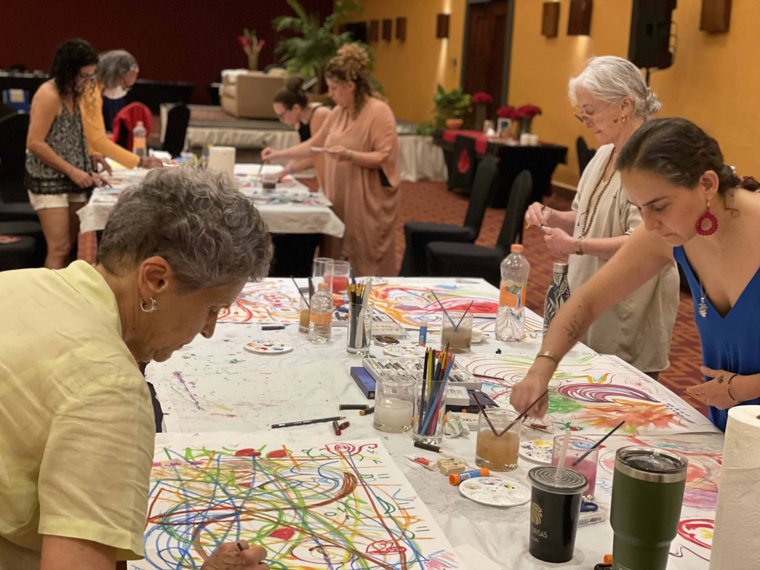 Group of women participating in a painting expressive arts activity at a workshop, with colorful abstract artwork on the tables.