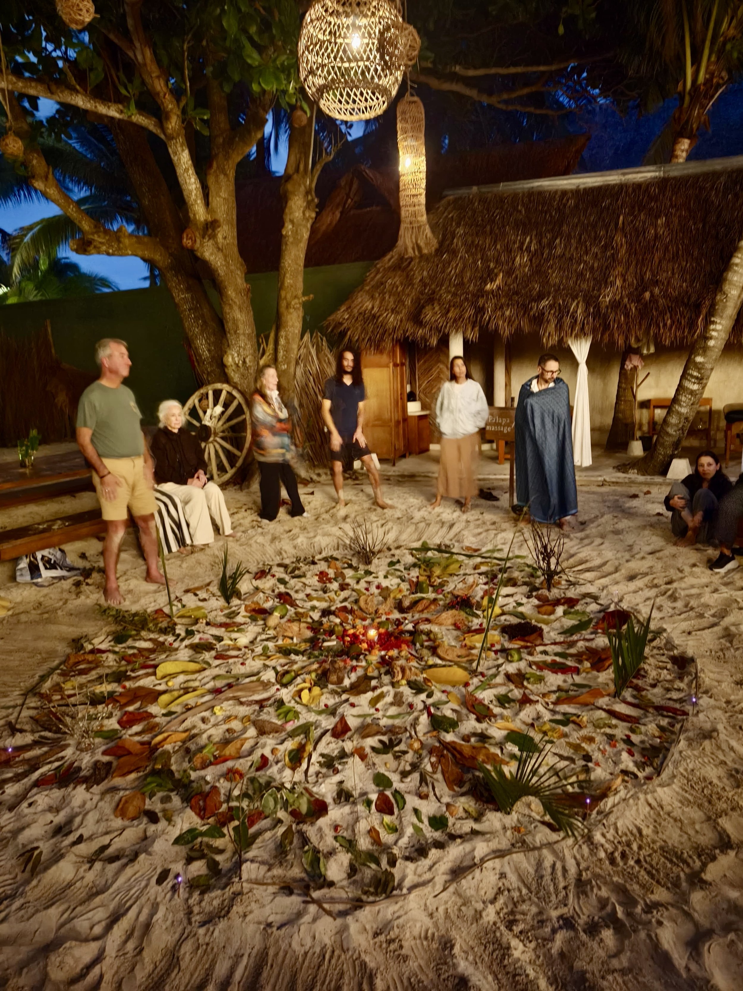 Group of people standing and sitting around a sand mandala with flowers and candles, on a beach at dusk, with tropical trees and a thatched roof hut in the background.