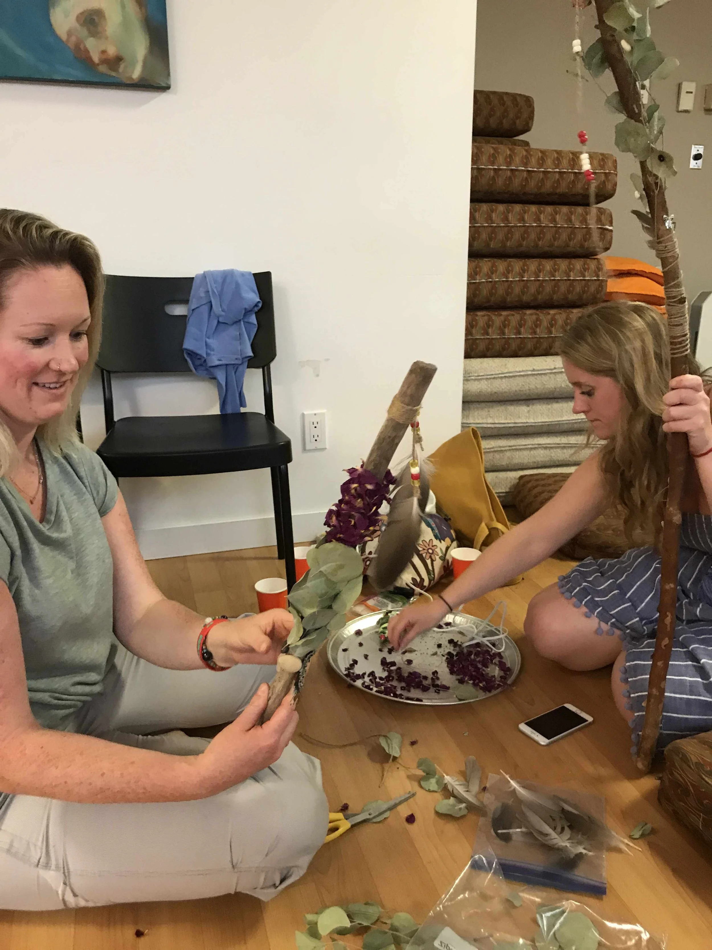 Two women sitting on the floor and crafting a decorative tree using branches, leaves, and flowers. One woman is holding a branch with leaves, and the other is reaching for flowers on a tray. There are crafting supplies scattered on the floor.