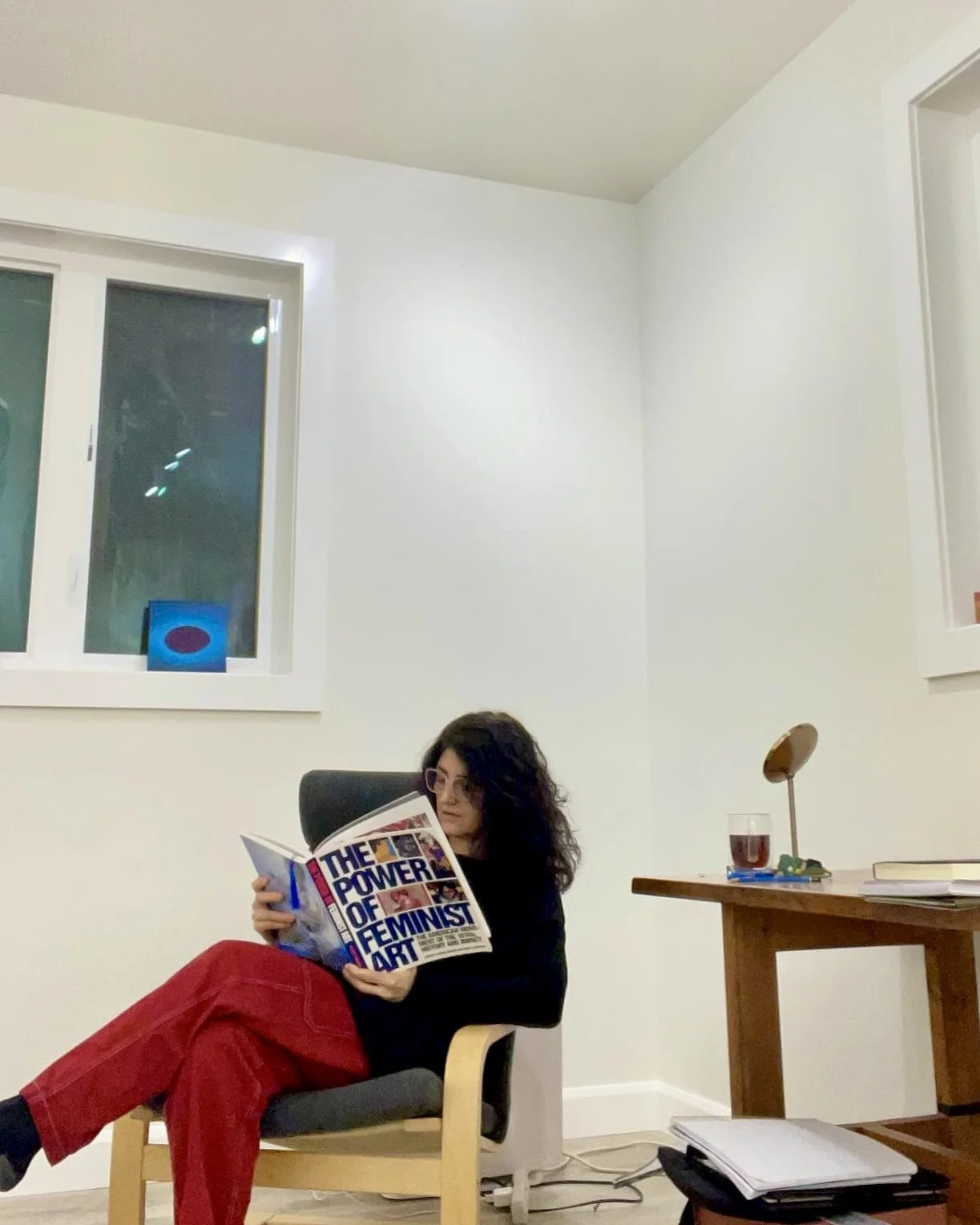 A contemporary Artist with curly hair reading a book titled 'The Power of Feminist Art' while sitting in a chair in a minimalistic room with a window, a desk with books, and a glass of red wine.