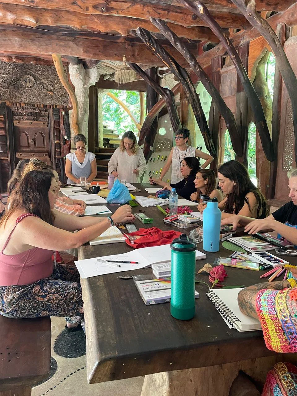 A group of people sitting around a large table doing creative work, in a room with nature-inspired decor, including wood and plant elements, engaged in a meeting or workshop.