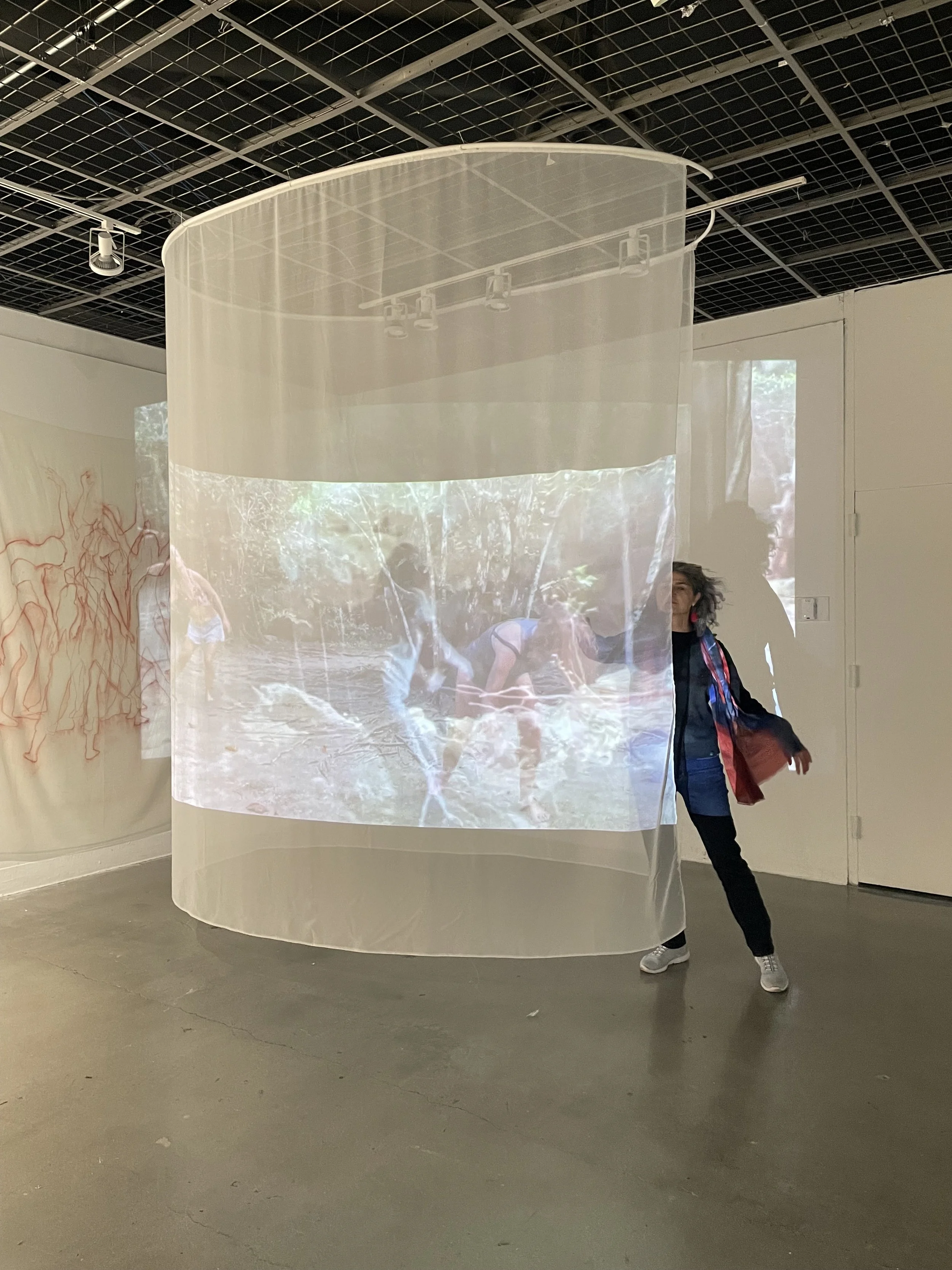 Person standing next to a large, curved, semi-transparent projection screen displaying an outdoor forest scene with people in it inside an art gallery.