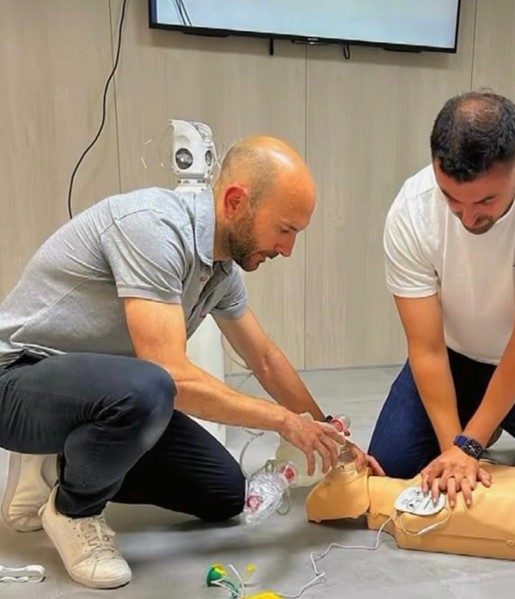 Dos hombres practican reanimación cardiopulmonar en un maniquí. Uno de ellos está usando un equipo de resucitación, y el otro lo ayuda en la simulación, en un ambiente interior.