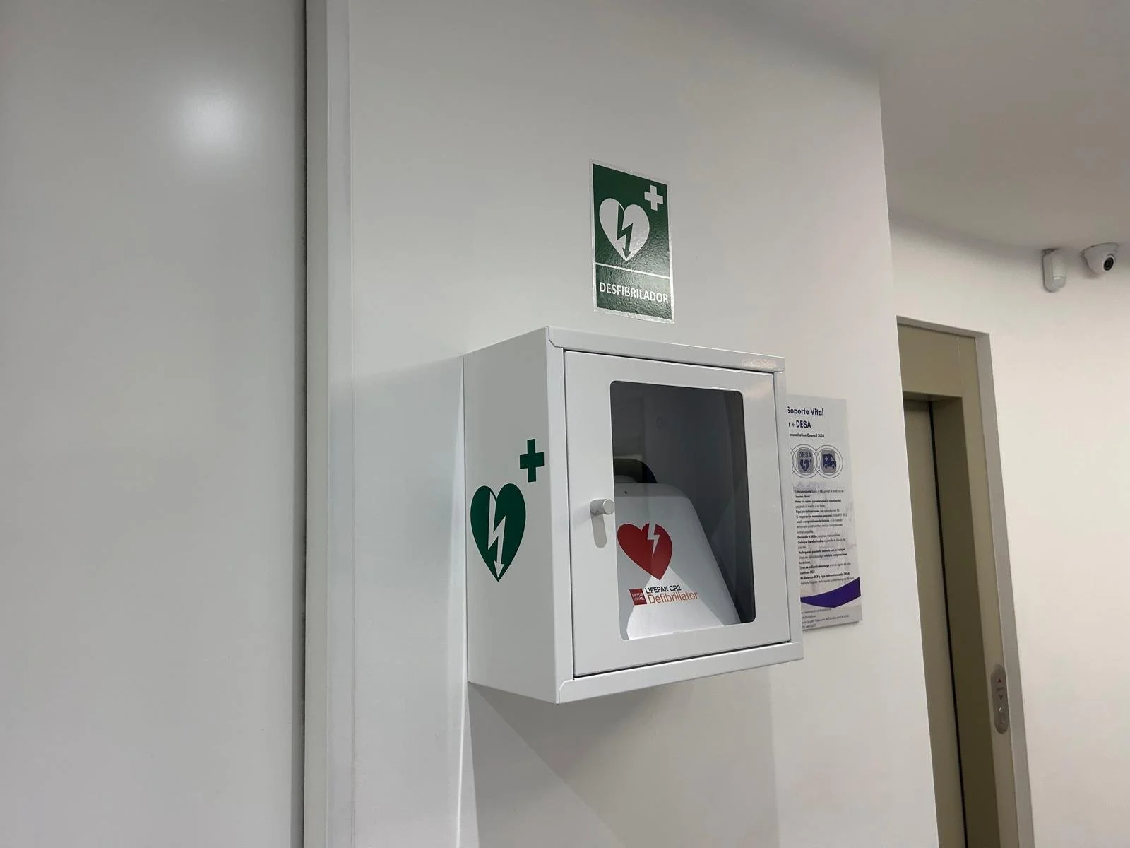 Caja de emergencia para desfibrilador en la pared de un pasillo, con señales verdes y un símbolo de corazón y un rayo, en un ambiente interior