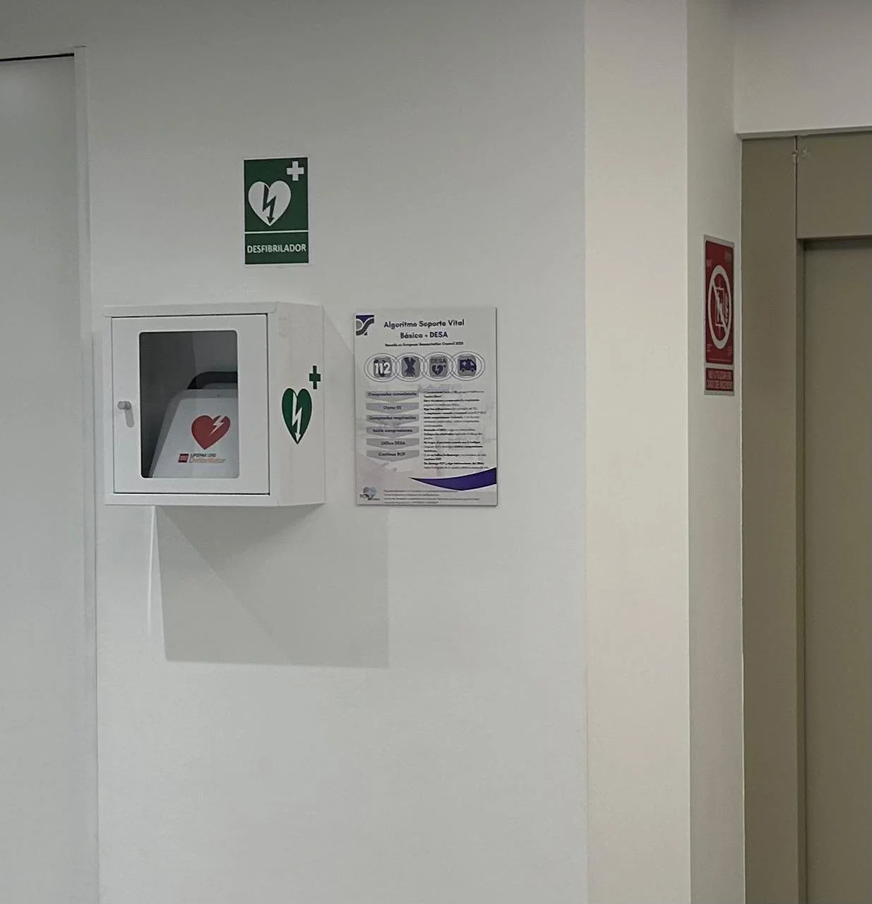 Casco de desfibrilador en pared blanca, señal de emergencia con corazón y rayo, cartel instructivo al lado.