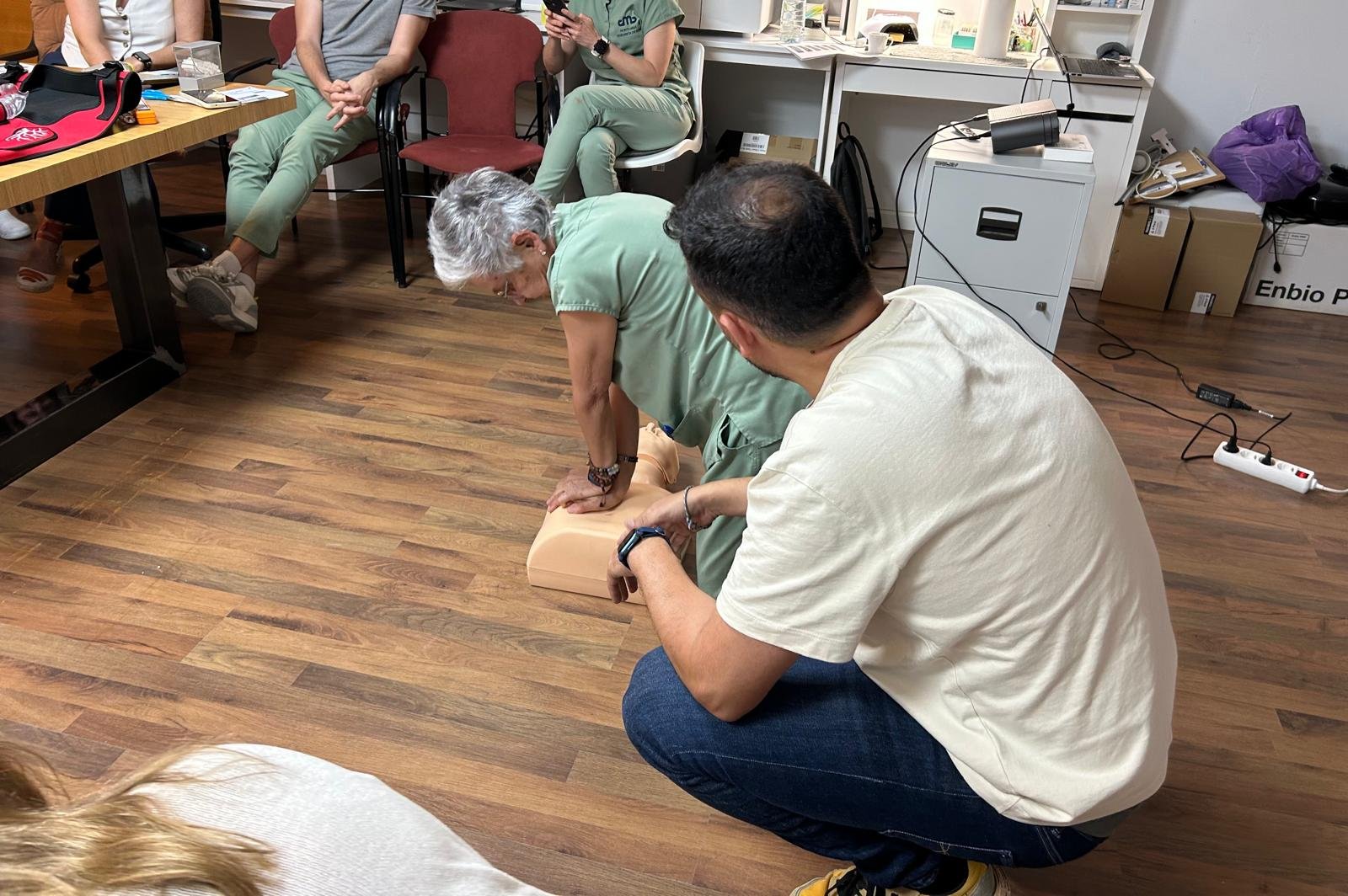 Persona realizando maniobra de reanimación en un torso de entrenamiento, mientras otra persona lo supervisa, en un espacio de oficina.