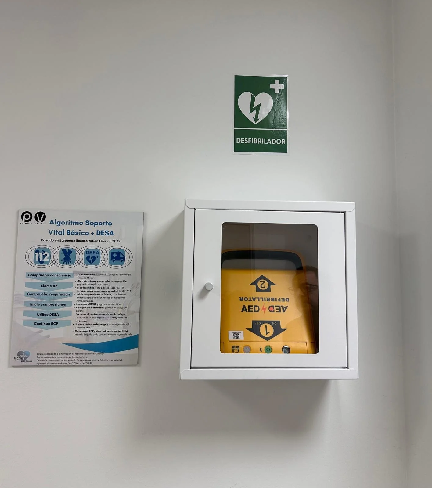 Altavoz de resucitación con instrucciones y un dispensador de desfibrilador automático externo (DEA) en la pared, con un cartel verde de 'Desfibrilador' y un símbolo de corazón con relámpago.