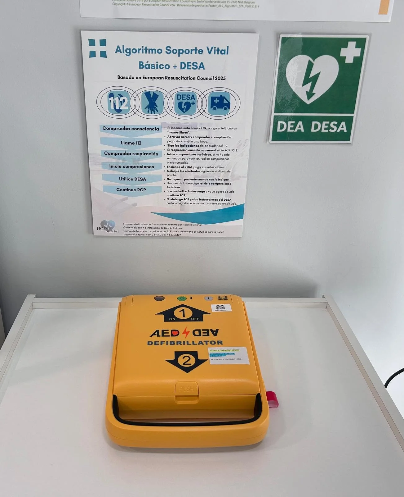 Un desfibrilador amarillo sobre una mesa, con instrucciones de uso y señalización de emergencia en la pared detrás, incluyendo un cartel de Algoritmo Soporte Vital Básico y DESA/DESA, y un signo verde con cruz blanca.