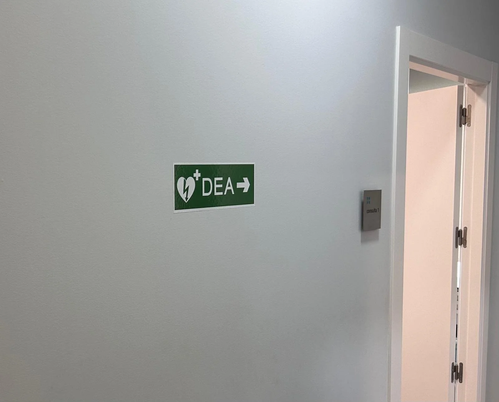 Señal de emergencia en pared blanca que indica DEA con flecha hacia la derecha, junto a una puerta abierta que revela un pasillo y una pared interior.