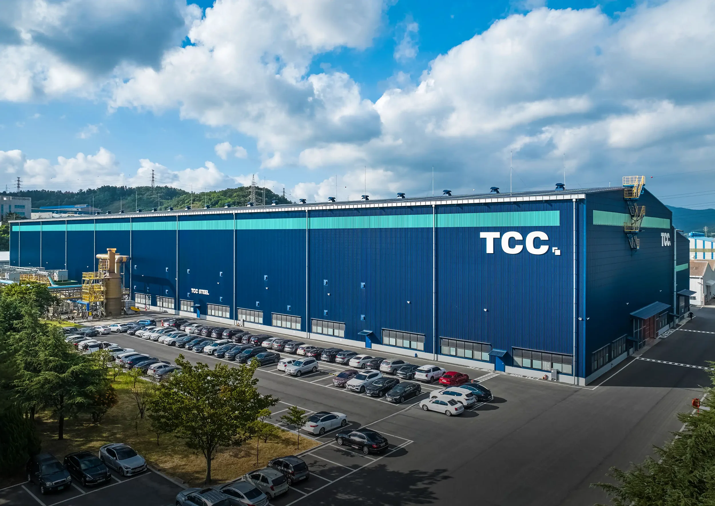 Large TCC steel manufacturing plant exterior with industrial facility, parking lot, and blue warehouse building under a clear sky