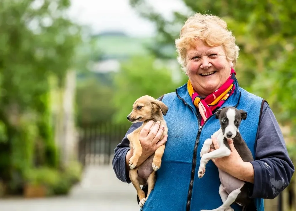 A smiling elderly woman holding two small puppies, one tan and one black and white, outdoors with green foliage in the background.