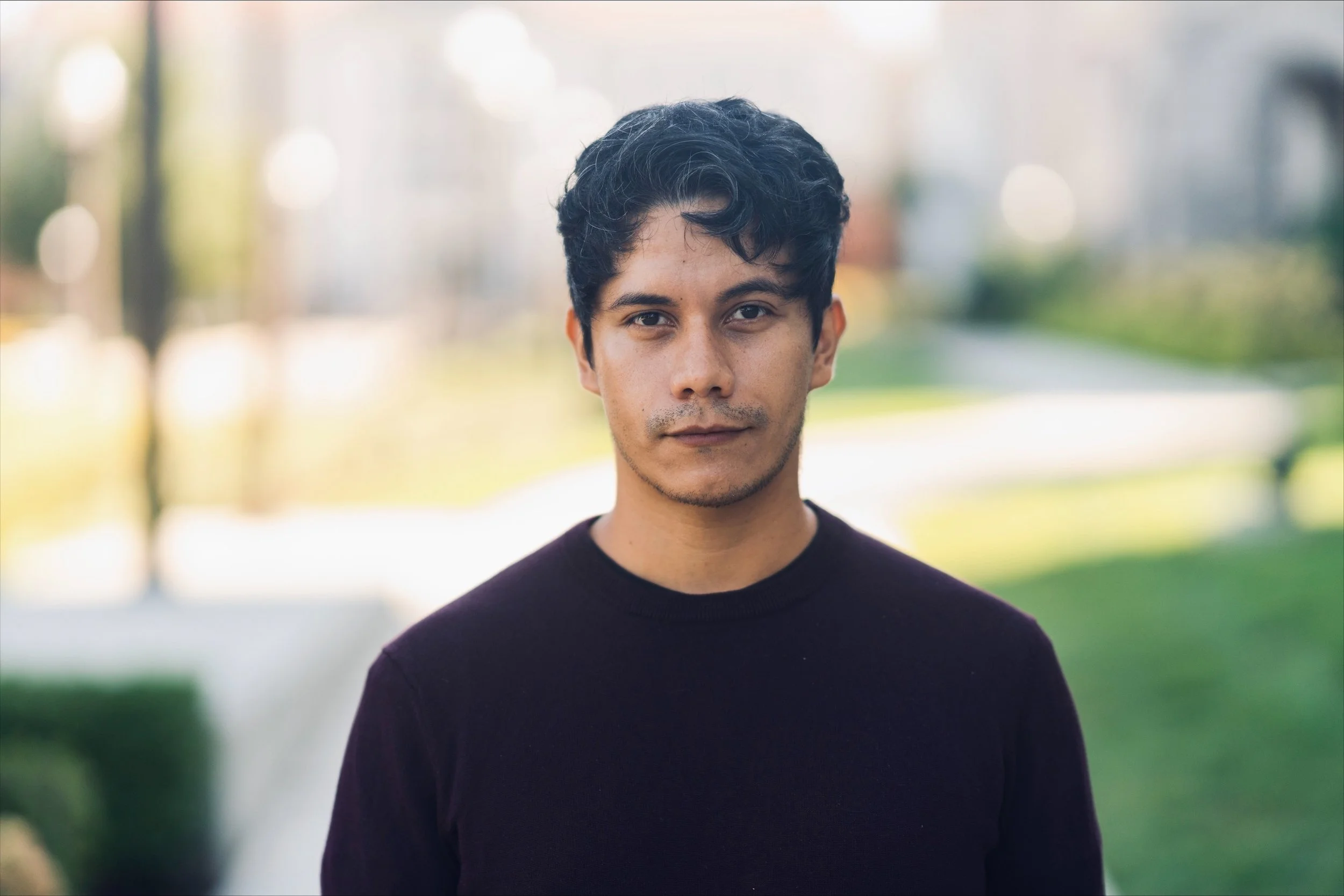 Young man with dark curly hair and light brown skin standing outdoors in a park, wearing a black shirt, with a blurred green and light background.