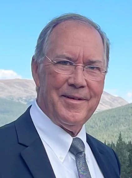 Headshot of a middle-aged man with glasses, wearing a dark suit, white shirt, and patterned tie, standing outdoors with mountains and a clear blue sky in the background.