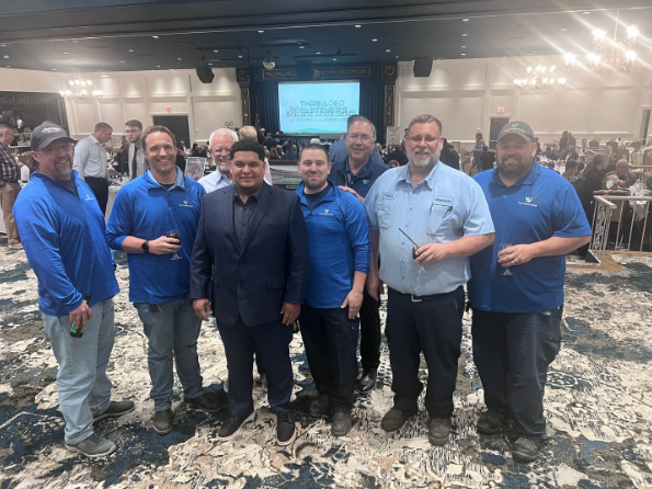 Group of seven men standing together at a conference, some in blue shirts, one in a suit, in a large banquet hall with tables and a stage in the background.