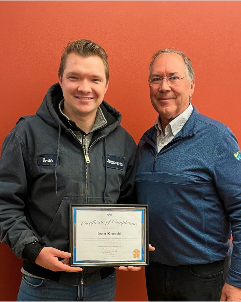 Two men standing next to each other, smiling, one holding a certificate of completion, against an orange wall.