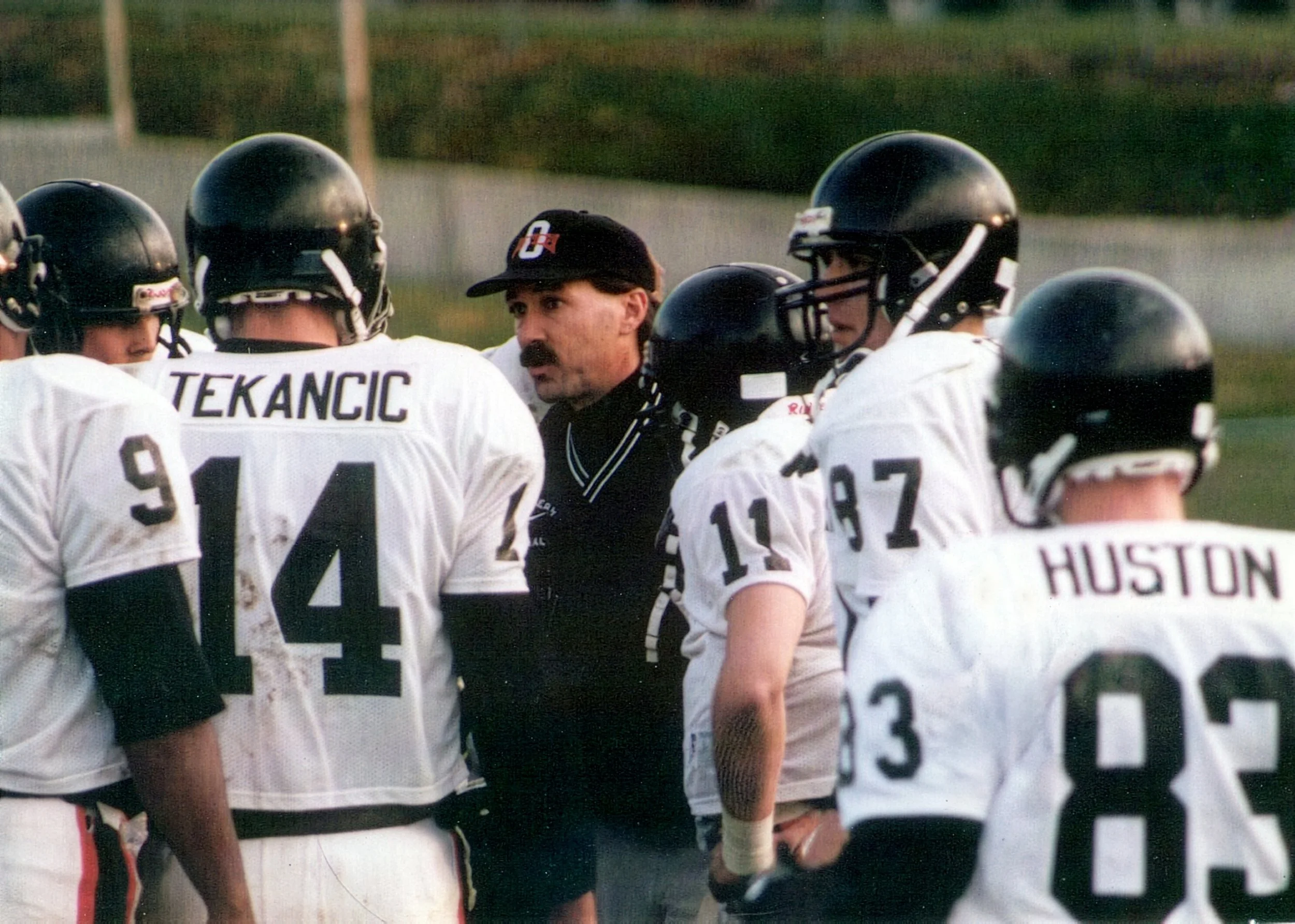 Football coach giving instructions to players during a game, players in white jerseys with black helmets, some with visible numbers and names, on a grassy field.