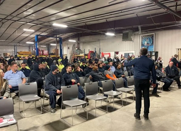 Group of people attending a presentation or meeting inside a large industrial warehouse.