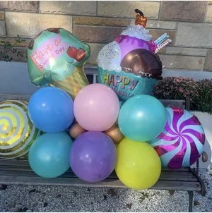 Colorful birthday balloons including cupcakes, candies, and a 'Happy Birthday' message, arranged outdoors on a bench.