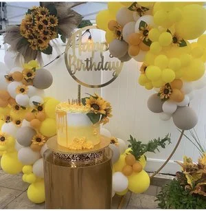 Birthday cake decorated with sunflowers and yellow accents, surrounded by balloon arch in yellow, white, gray, and orange, with a 'Happy Birthday' sign