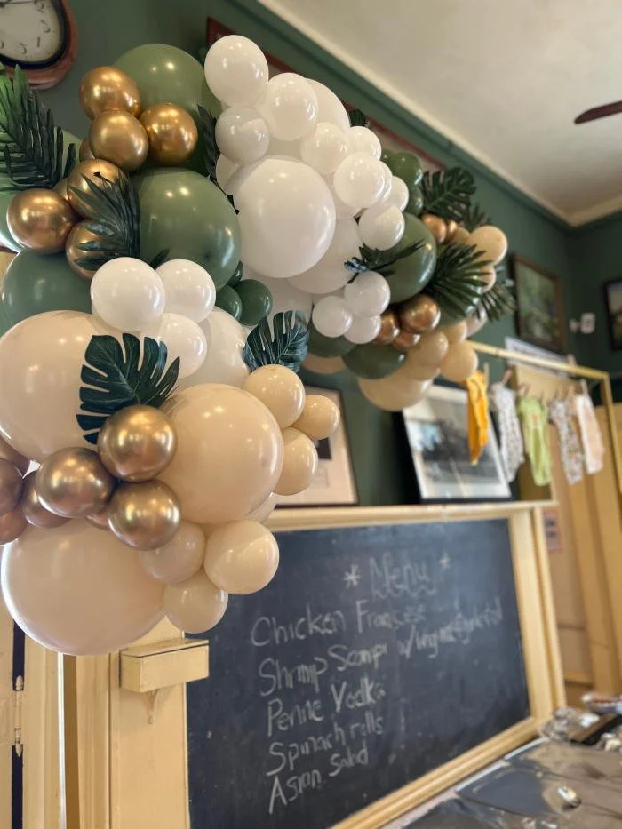 Decorative balloon garland in white, green, cream, and gold with tropical leaves, set against a restaurant interior with a chalkboard menu.