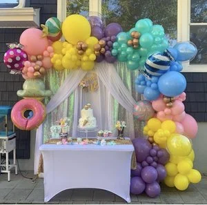 Colorful balloon arch with pastel and bright balloons framing a dessert table with a cake and cupcakes for a celebration.