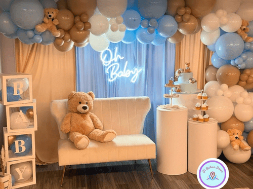 Decorated baby shower area with beige, white, blue, and brown balloons, a white loveseat with a teddy bear, a tiered dessert stand, and a neon sign that reads 'Oh Baby'.