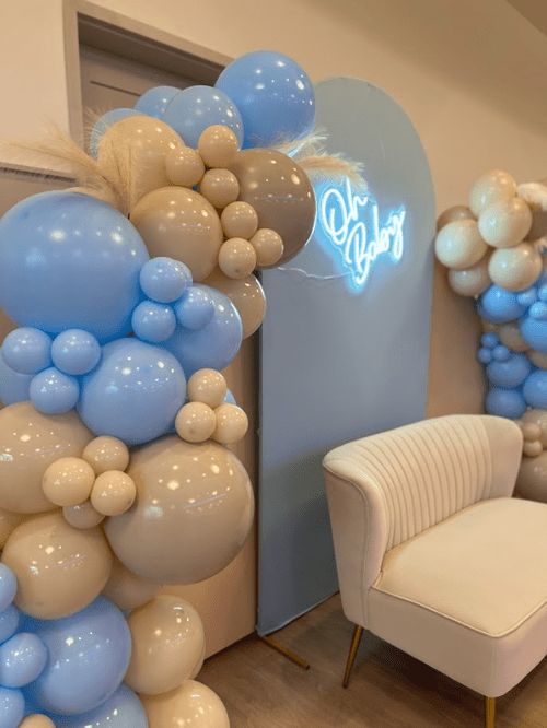 Balloon arrangements in blue, white, and beige surrounding a light blue backdrop with a neon sign that reads 'Oh Baby' and a cream-colored upholstered armchair.