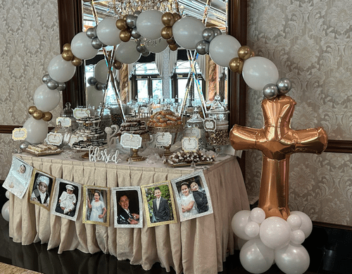A dessert table decorated for a celebration, featuring an arch made of gold, silver, and white balloons, with a large balloon cross at the side. The table has various treats and a sign that says 'blessed' in the center.