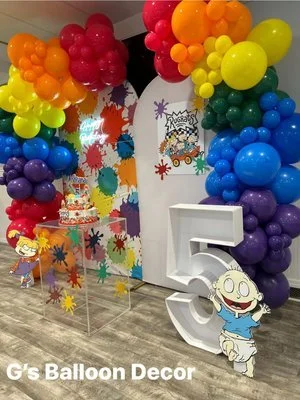 Colorful balloon arch with rainbow colors and a birthday party setup featuring a large number five, cartoon character cutouts, and a birthday cake.
