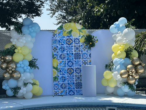Decorative backdrop with blue and yellow tile-patterned panels, surrounded by balloon arrangements in white, blue, yellow, and gold, with greenery accents.