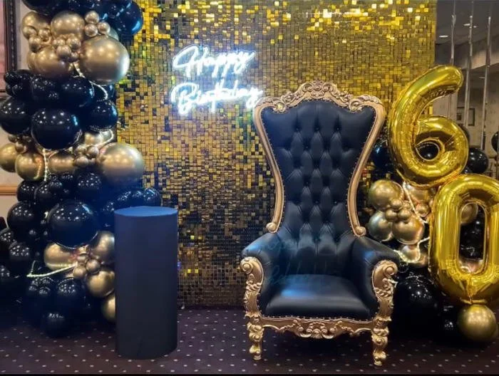 Decorative birthday celebration setup with a blue and gold theme, featuring a large ornate black chair, black and gold balloons, a gold number 60 balloon, a "Happy Birthday" neon sign, and a golden sequin backdrop.