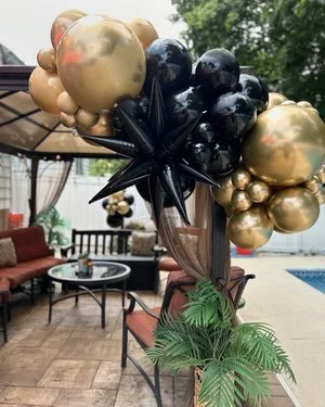 Balloon centerpiece with gold and black balloons and black star-shaped balloons, placed on a table with tropical leaves in an outdoor patio setting.