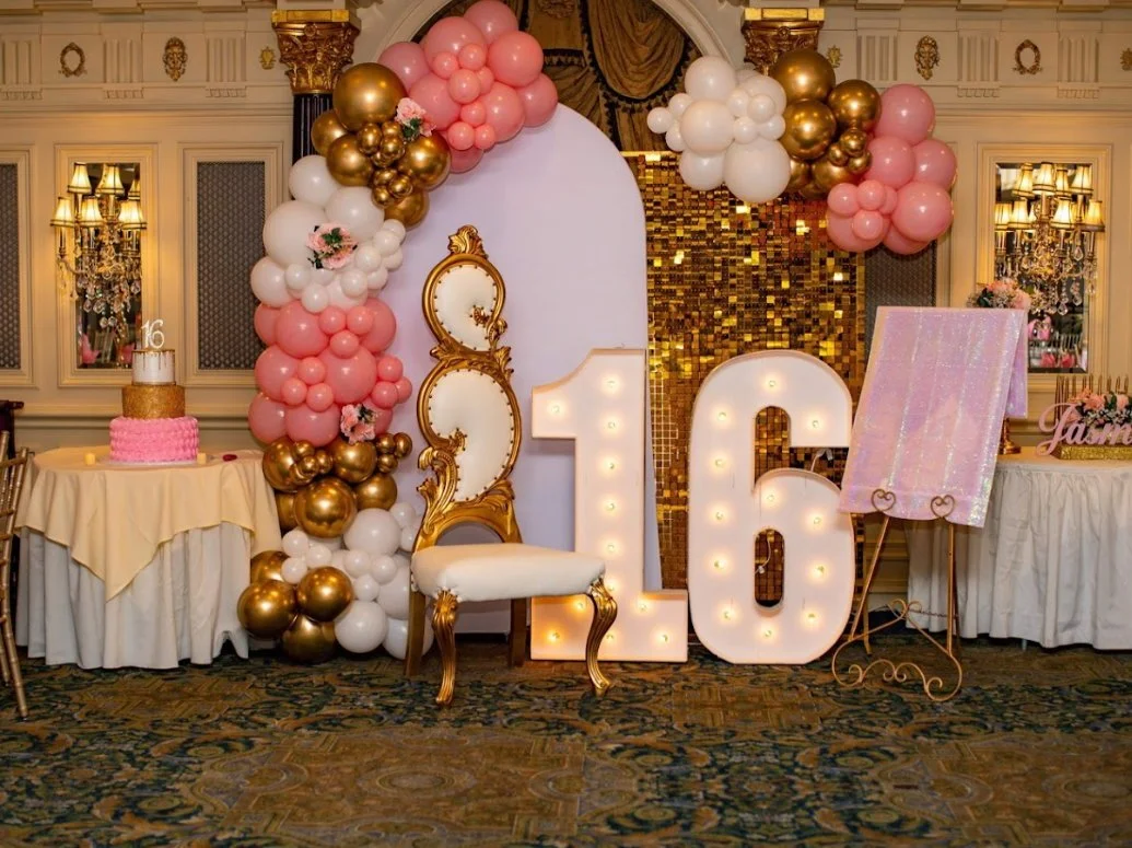 Decorative setup for a 16th birthday celebration with large illuminated numbers '16', pink, white, and gold balloons, a vintage gold-trimmed chair, a cake, and an easel with a pink canvas.
