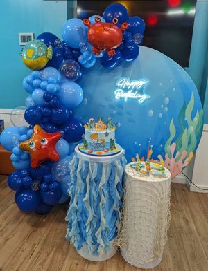 Sea-themed birthday decoration with blue balloons, a crab and starfish balloon sculptures, a cake with ocean decorations, and a neon sign saying 'Happy Birthday'.