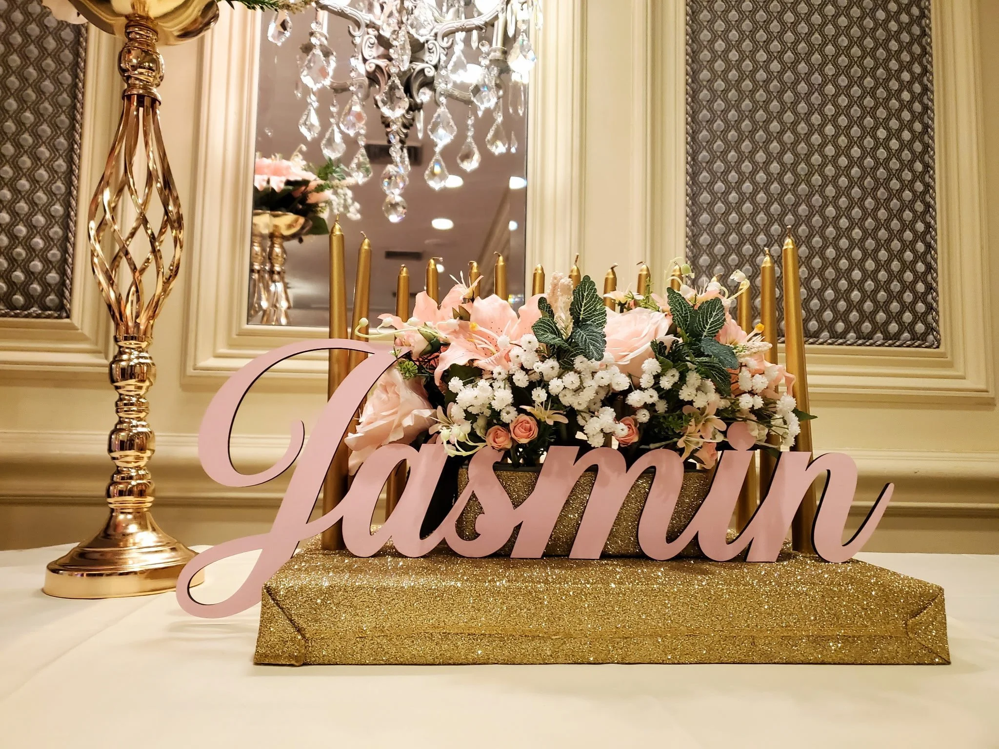 Table centerpiece with pink flowers, a pink 'Jasmin' sign, and gold candles, set against a mirror and ornate decor in the background.