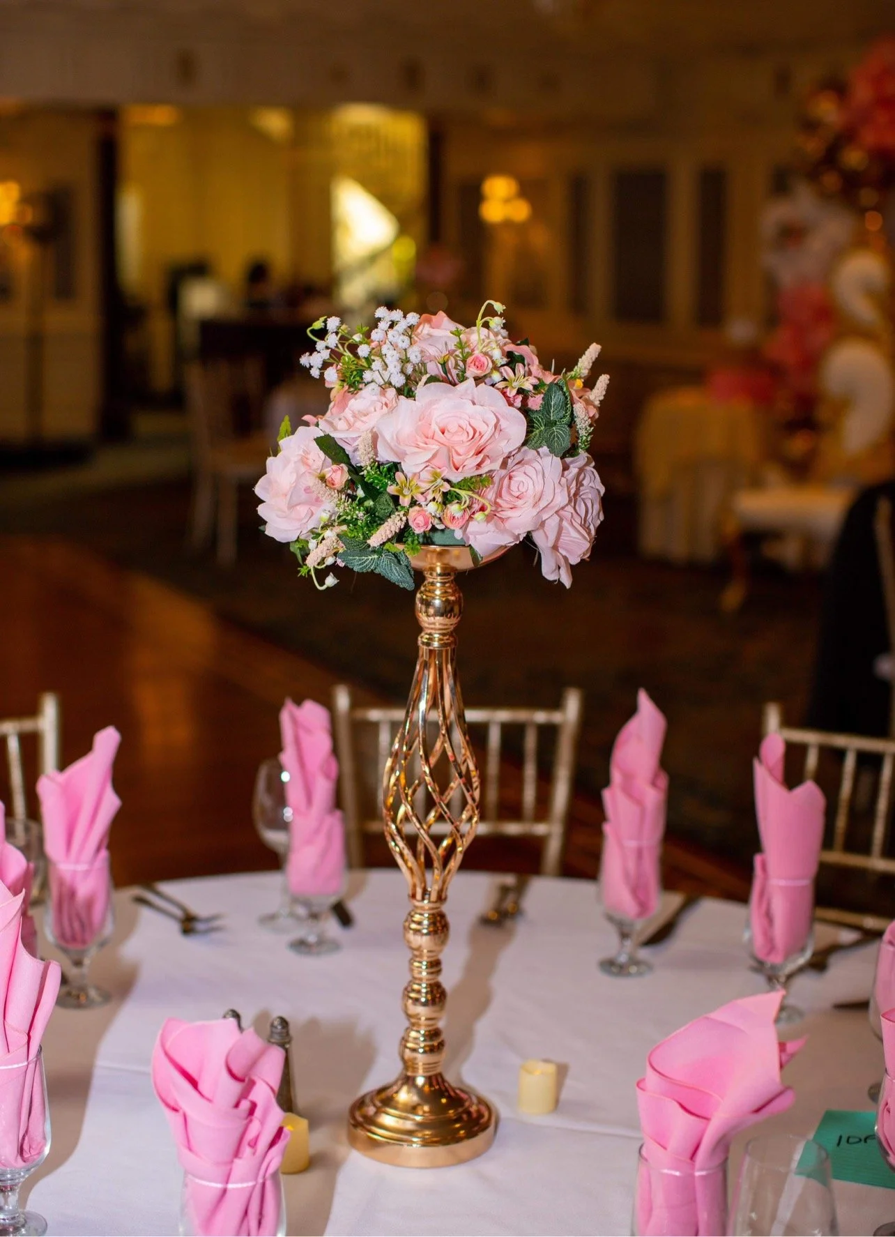 A floral centerpiece with pink roses on a tall gold stand, surrounded by pink napkins folded in glasses on a round dining table.