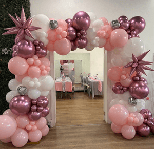 Balloon arch with pink, white, and purple balloons, decorated with star-shaped balloons, framing the entrance to a decorated party room with tables and chairs.