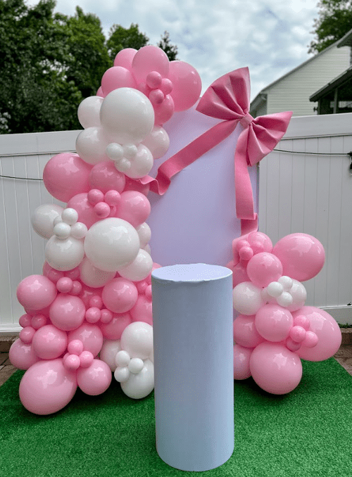 Pink and white balloon arrangement with a pink ribbon bow and a white backdrop, set in an outdoor space.