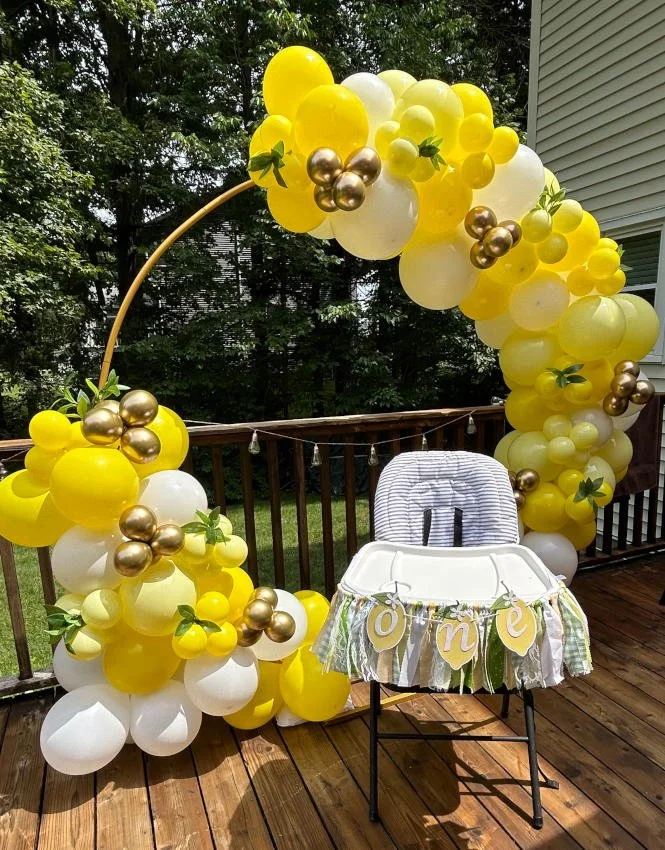 Decorative balloon arch in yellow, white, and gold colors on a wooden deck, with a high chair and a banner spelling 'one' in front of the chair.