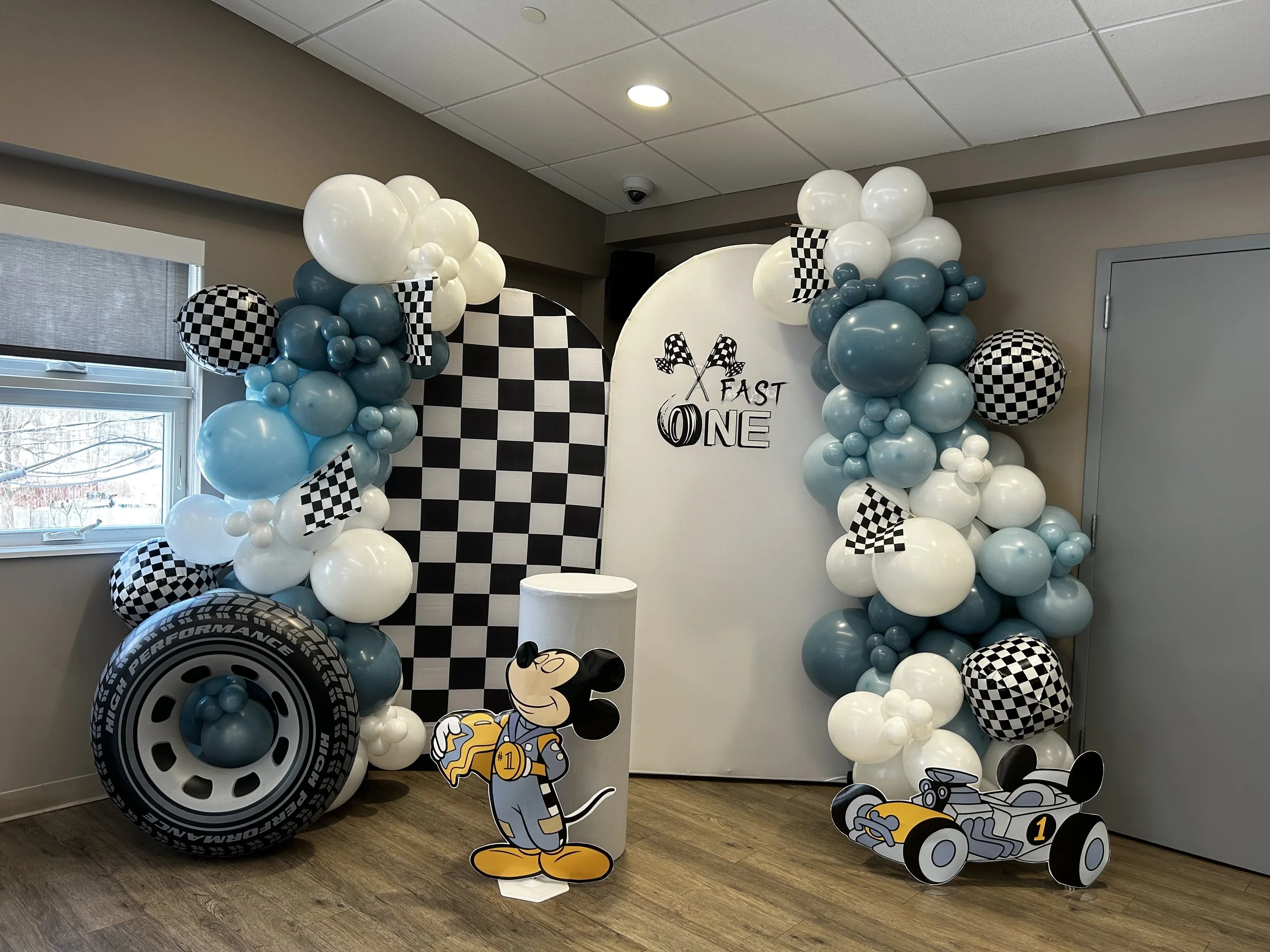 Decorative racing-themed backdrop with checkered flags, balloons in black, white, and blue, a tire prop, and cartoon Mickey Mouse and race car cutouts, celebrating a first birthday.