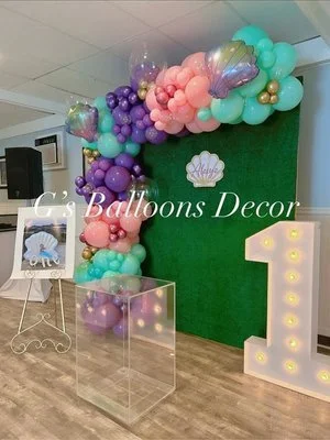 Balloon decoration with pastel and purple balloons, a green backdrop, a large illuminated number one.