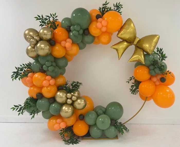 Decorative wreath made of balloons in orange, green, gold, peach, and black colors, accented with green leaves and a large gold bow.