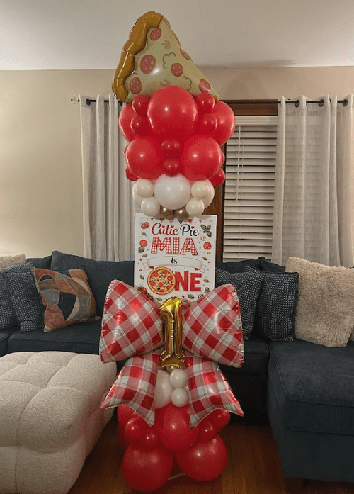Decorative balloon arrangement for a first birthday celebration with a cake-shaped balloon at the top, red, white, and gold balloons.