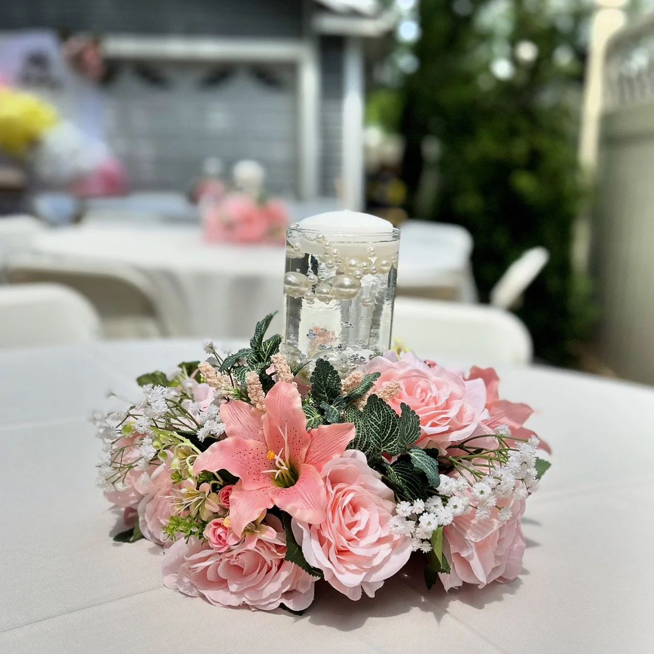 A floral centerpiece with pink roses, lilies, and other small flowers surrounding a candle in a glass holder filled with water and floating bubbles, on a white tablecloth.
