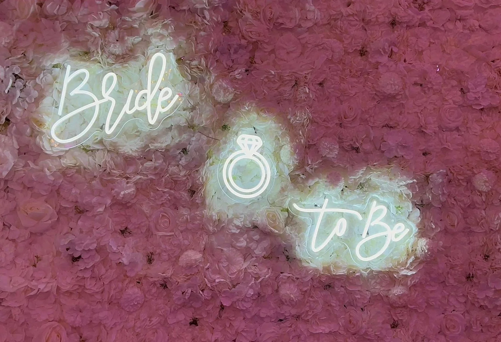 Neon sign with the words 'Bride to Be' illuminated against a backdrop of pink and white flowers.